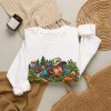 White crewneck sweatshirt with "In the Bushes Finding Myself" disc golf graphic, lifestyle flat-lay