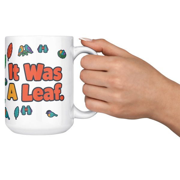 "I watched it for ten minutes. It was a leaf." Birding 15oz mug, raised in hand 15 oz white ceramic birding mug, "I watched it for ten minutes" on one side and "It was a leaf" on the other, with birds, binoculars and leaves, raised in hand