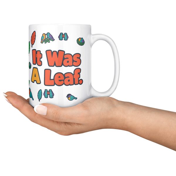 "I watched it for ten minutes. It was a leaf." Birding 15oz mug, held in an open hand 15 oz white ceramic birding mug, "I watched it for ten minutes" on one side and "It was a leaf" on the other, with birds, binoculars and leaves, held in an open hand
